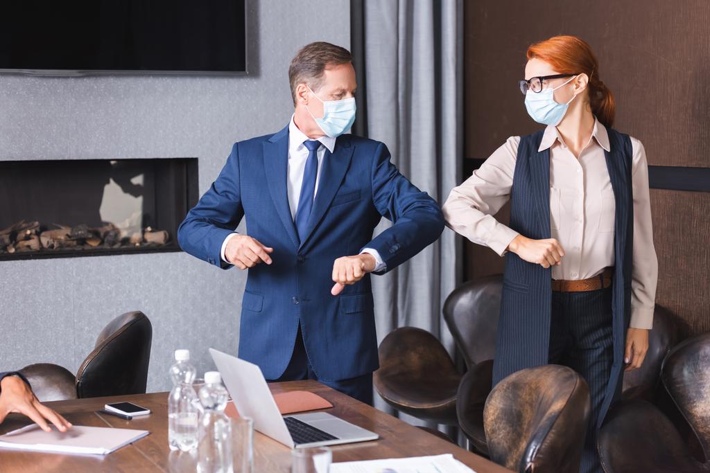 Femme d'affaires rousse et homme d'affaires en masques médicaux se saluant les uns les autres avec les coudes dans la salle de conférence - Photo, image