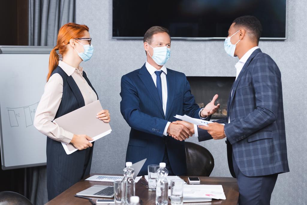Multikulturelle Geschäftsleute in medizinischen Masken schütteln einander die Hände, während sie neben Geschäftsfrau im Sitzungssaal stehen - Foto, Bild