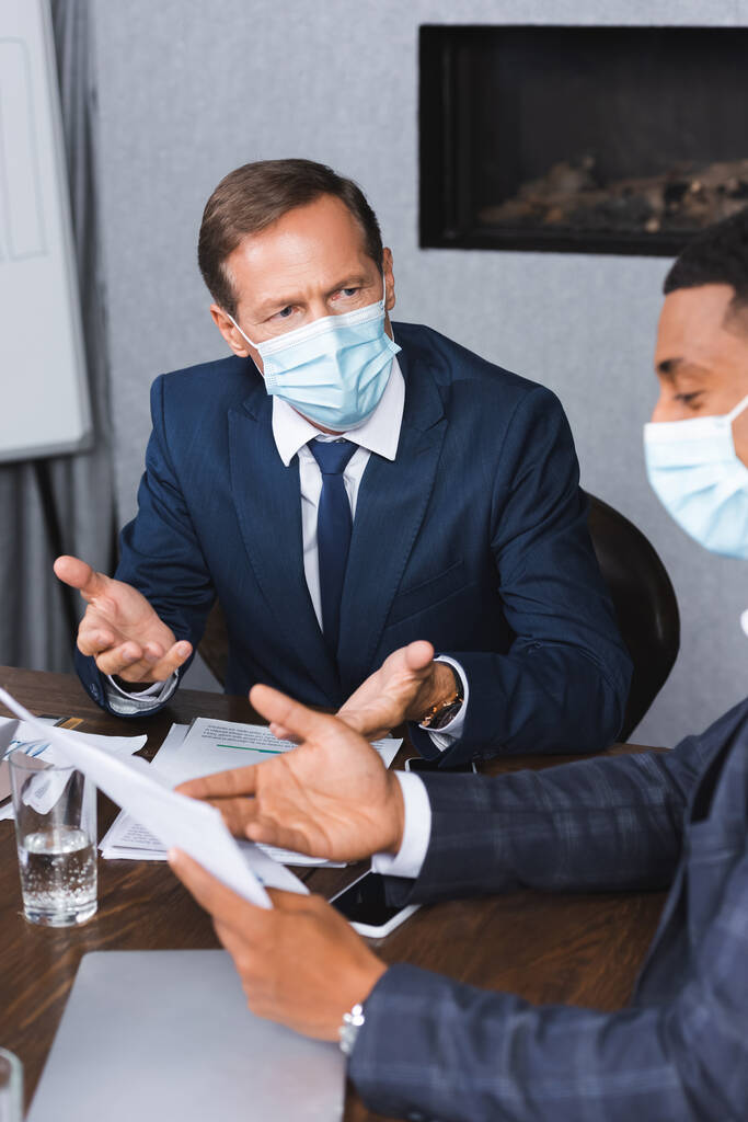 Businessman in medical mask gesturing while looking at african american colleague with paper at workplace - Photo, Image