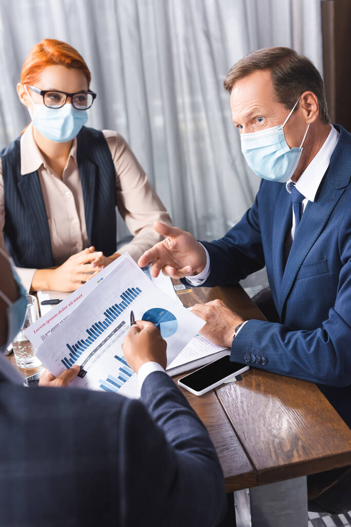 Empresario en máscara médica mirando a colega afroamericano mostrando papeles con gráficos en primer plano borroso - Foto, Imagen