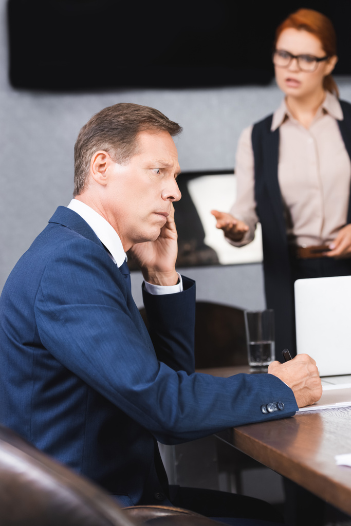 Τρομαγμένος επιχειρηματίας κάθεται στο χώρο εργασίας με θολή θυμωμένος εκτελεστικό στο παρασκήνιο - Φωτογραφία, εικόνα