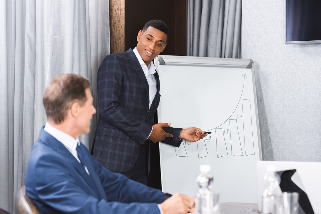 Улыбающийся африканский американский бизнесмен, указывая ручкой на график на флипчарте и глядя на размытого коллегу на переднем плане - Фото, изображение