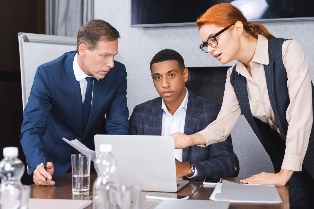 Серьезный африканский американский бизнесмен смотрит в камеру рядом с ноутбуком и коллегами, стоящими рядом на размытом переднем плане - Фото, изображение