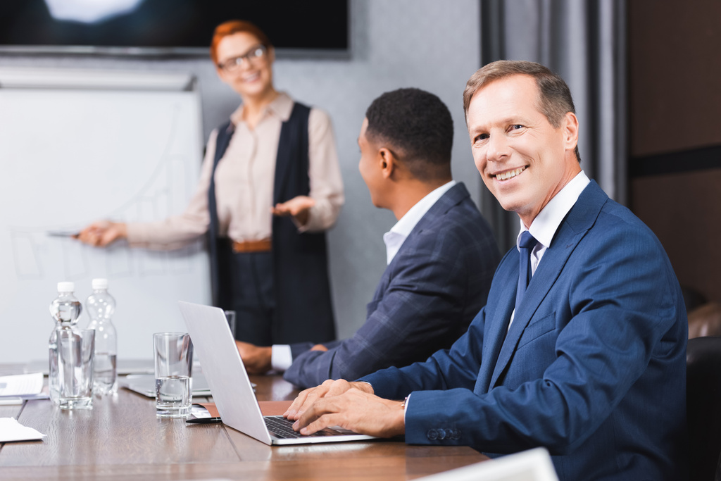 Smiling businessman looking at camera while typing on laptop with blurred multicultural colleagues on background - Photo, Image
