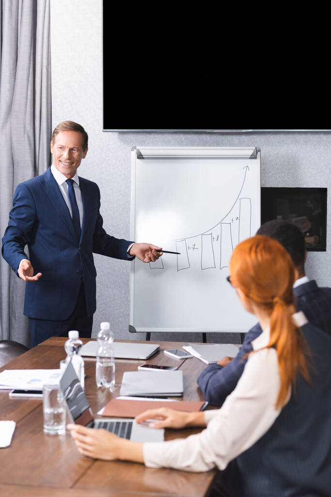 Smiling executive pointing with pen at graph on flipchart with blurred multicultural colleagues on foreground in meeting room - Photo, Image