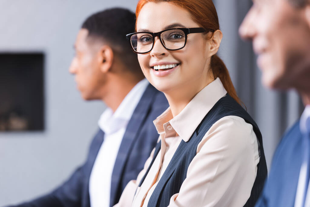Smiling redhead businesswoman in eyeglasses looking at camera with blurred african american colleague on background - Photo, Image