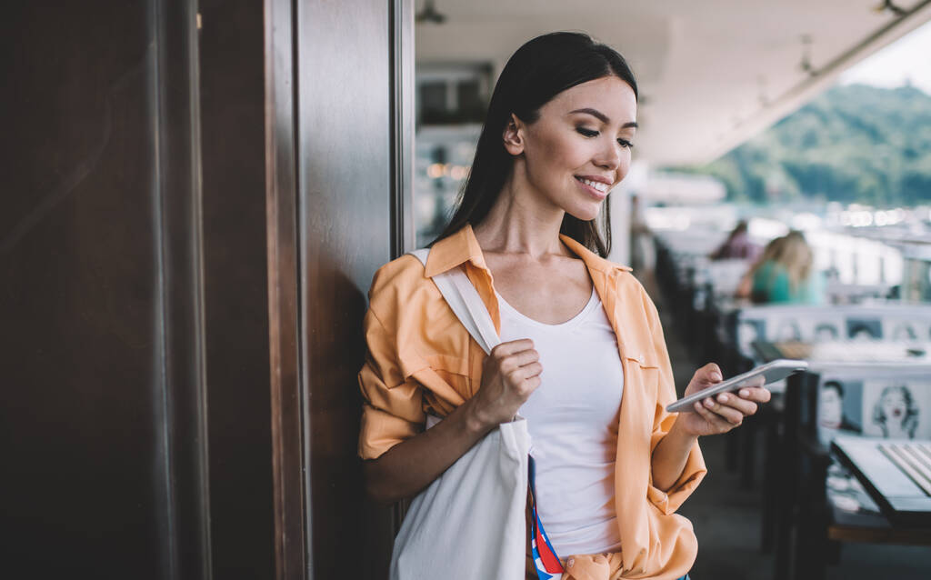 Junge zufriedene ethnische Frau in legerer Kleidung, die tagsüber auf der Terrasse eines Cafés steht und SMS aufs Handy sendet - Foto, Bild