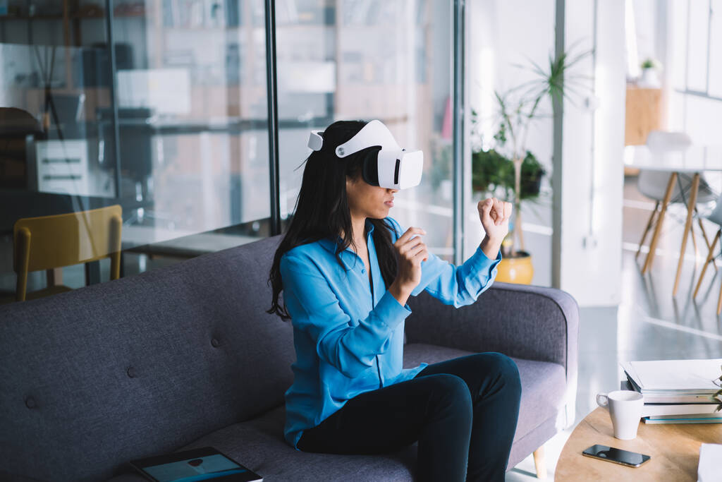 Vista lateral de una joven hembra enfocada con auriculares VR sentada en el sofá y haciendo gestos como sostener un objeto virtual en las manos - Foto, imagen