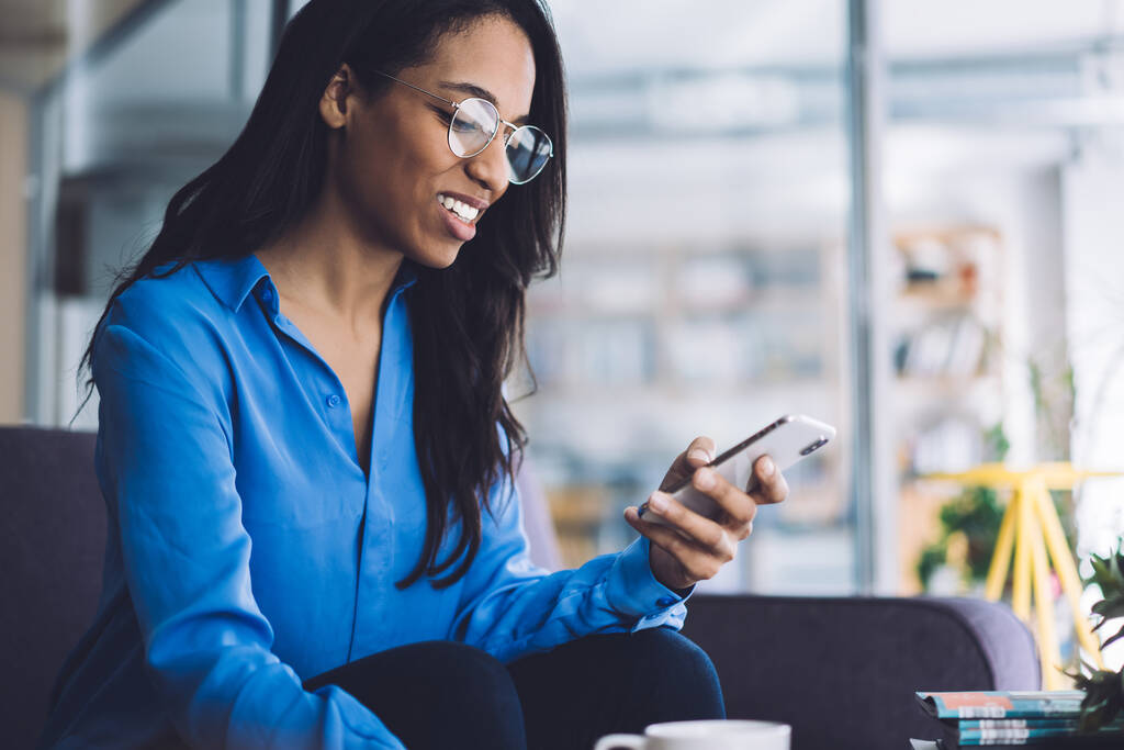 Usmívající se mladá Afroameričanka v ležérní oblečení a s brýlemi sedí na gauči procházení mobilní a pití šálek čaje - Fotografie, Obrázek