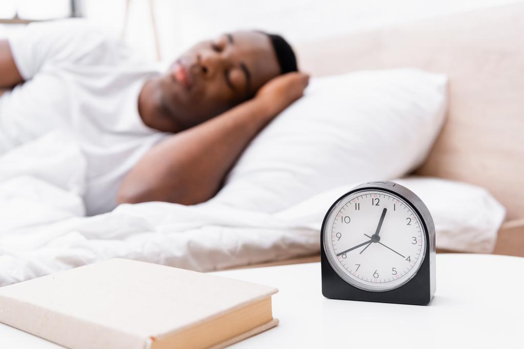 Zegar i książka na stoliku nocnym w pobliżu Afryki Amerykanin śpi na rozmazanym tle  - Zdjęcie, obraz