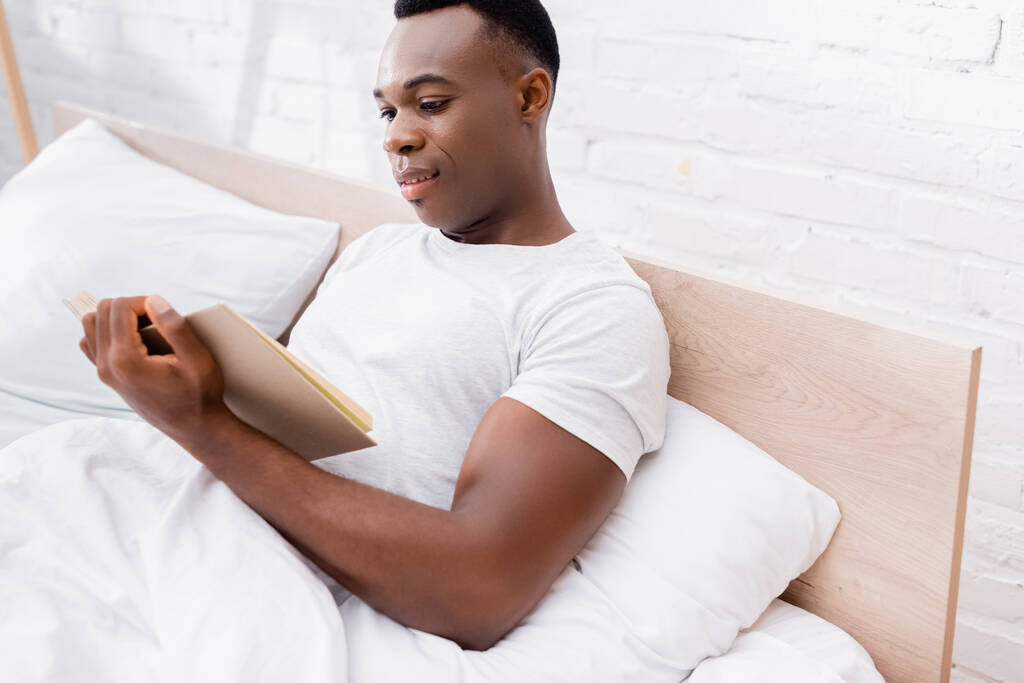 Usmívající se africký Američan čtení knihy na posteli s bílým lůžkoviny  - Fotografie, Obrázek
