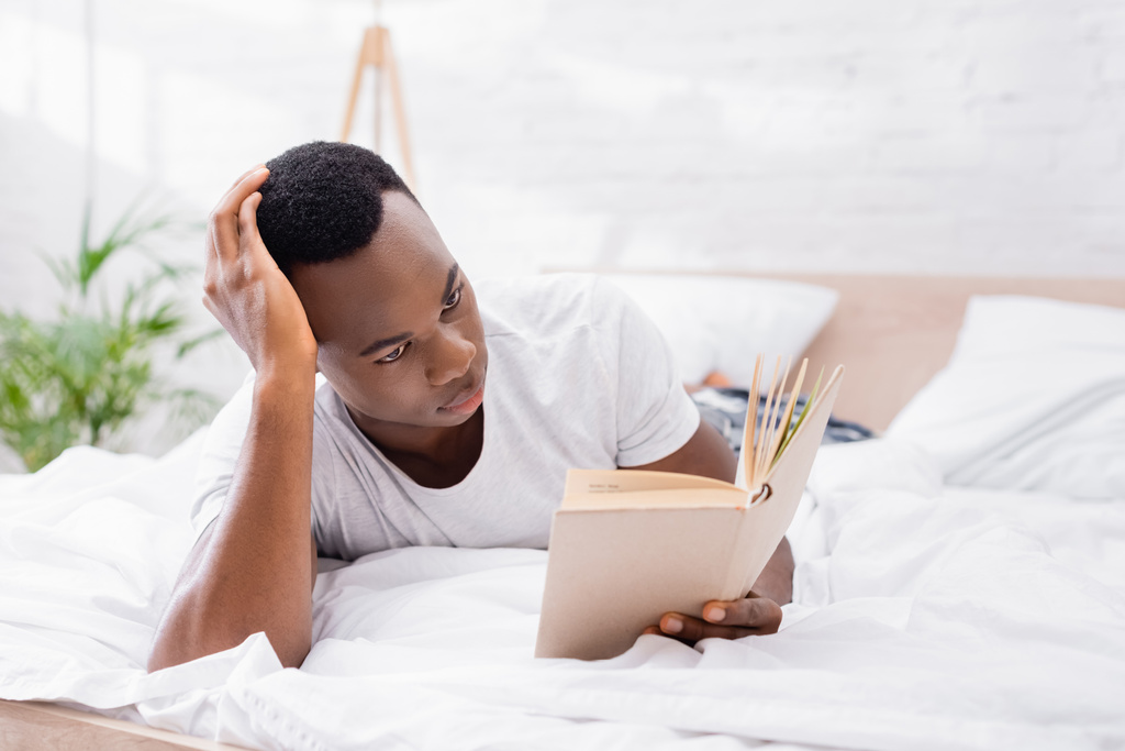 Hombre afroamericano enfocado leyendo libro en la cama en casa  - Foto, Imagen