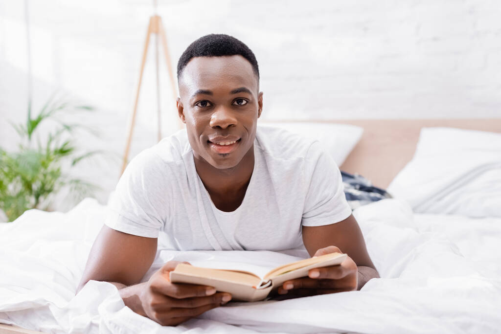Lächelnder afrikanisch-amerikanischer Mann blickt in die Kamera, während er Buch auf verschwommenem Vordergrund auf dem Bett hält  - Foto, Bild