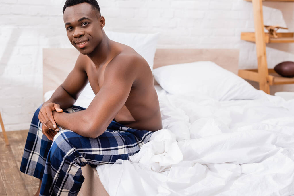 Póló nélküli afro-amerikai férfi ül az ágyon és mosolyog a kamerába  - Fotó, kép