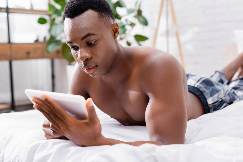 Hombre afroamericano sin camisa usando tableta digital en primer plano borroso en la cama  - Foto, imagen