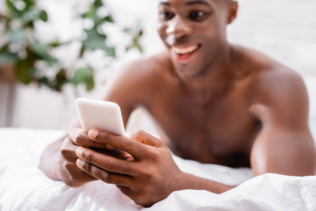 Smartphone dans les mains de l'homme afro-américain joyeux sur fond flou dans la chambre  - Photo, image
