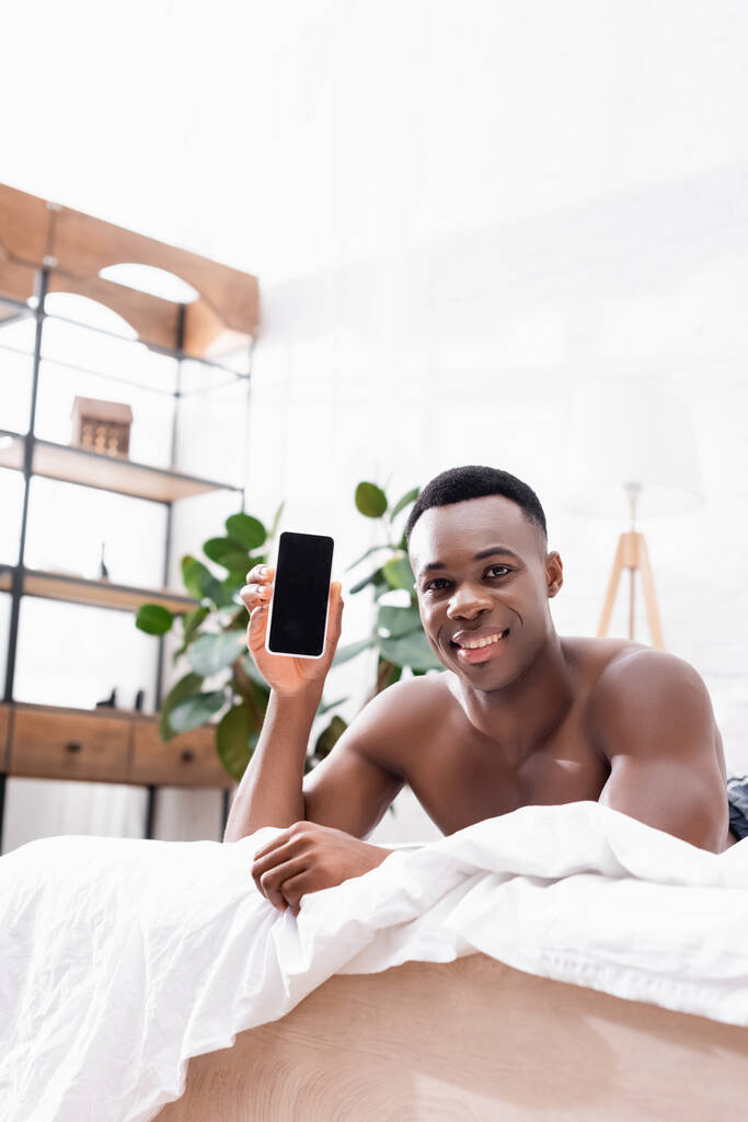 アフリカ系アメリカ人男性の笑顔とベッドの上に空白の画面でスマートフォンを示す  - 写真・画像