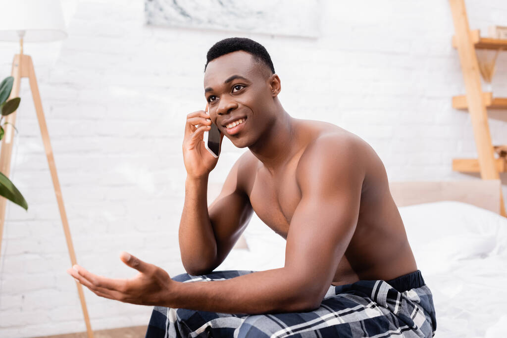 Shirtless Αφρικής Αμερικανός άνθρωπος χαμογελώντας, ενώ μιλάμε στο smartphone στην κρεβατοκάμαρα  - Φωτογραφία, εικόνα