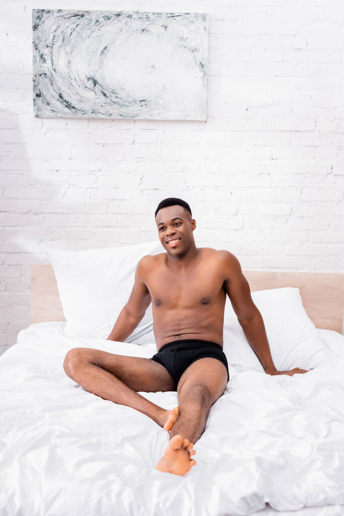 Веселый африканский американец в трусах, сидящий на кровати утром  - Фото, изображение