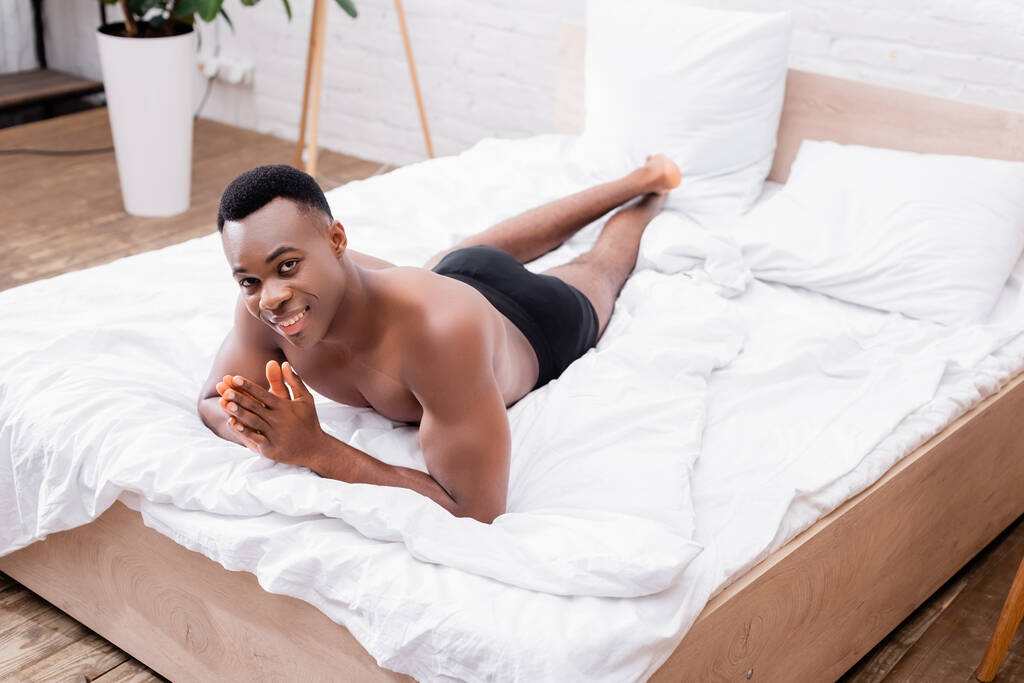 Sonriente afroamericano en calzoncillos acostado en la cama en casa  - Foto, imagen