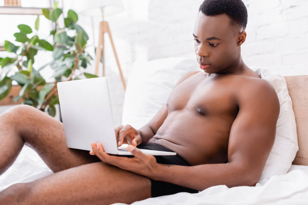 Африканский мужчина без рубашки использует ноутбук, когда лежит на кровати  - Фото, изображение