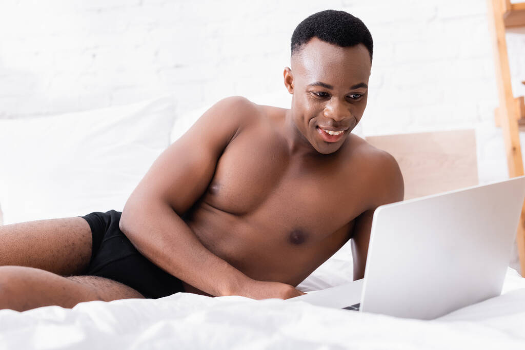 Uśmiechnięty Afroamerykanin w bieliźnie korzystający z laptopa na niewyraźnym pierwszym planie w łóżku  - Zdjęcie, obraz