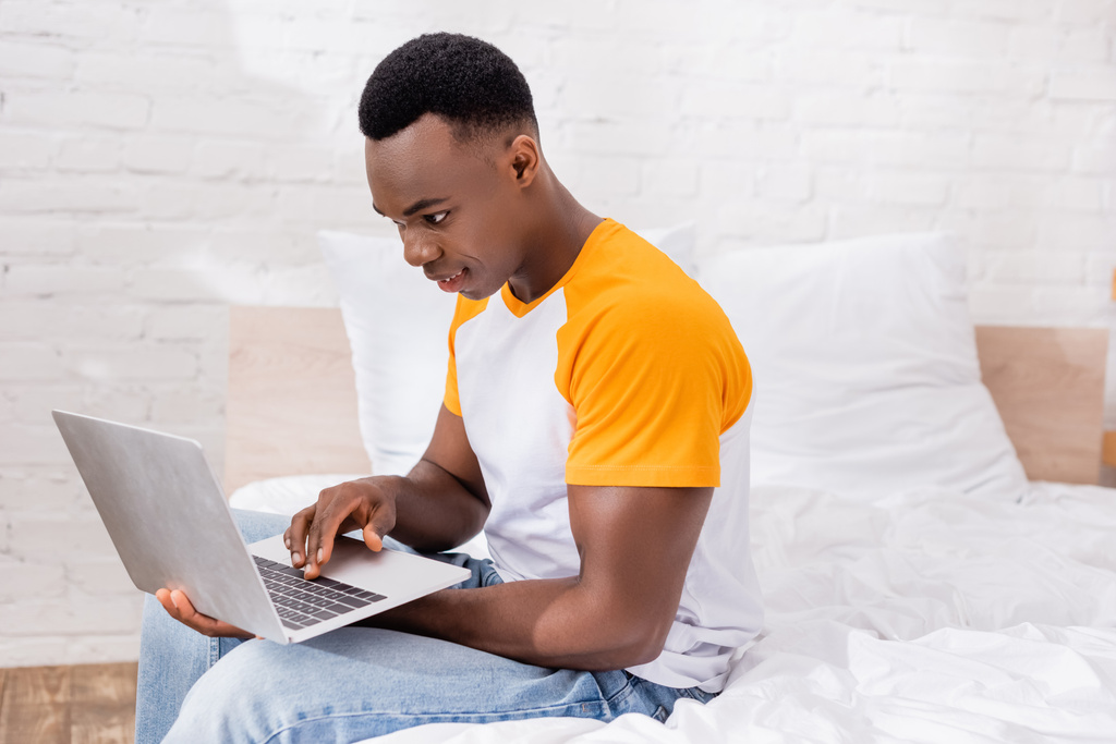Homem americano africano positivo usando laptop enquanto sentado na cama em casa  - Foto, Imagem