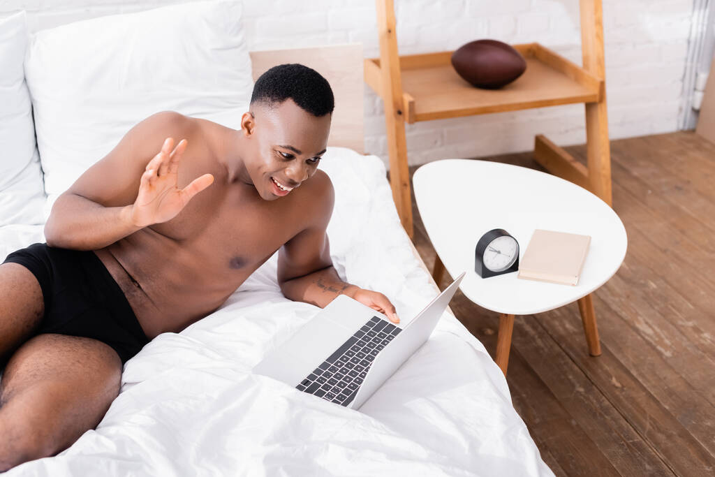 Cheerful shirtless african american man waving hand during video call on laptop near book and clock on coffee table  - Photo, Image