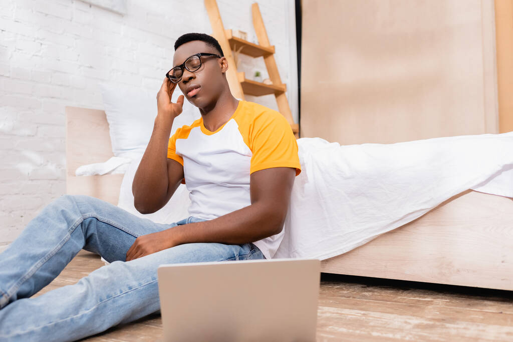 疲れアフリカ系アメリカ人男性で眼鏡で座っている近くのノートパソコンでぼやけた前景にベッドルーム  - 写真・画像