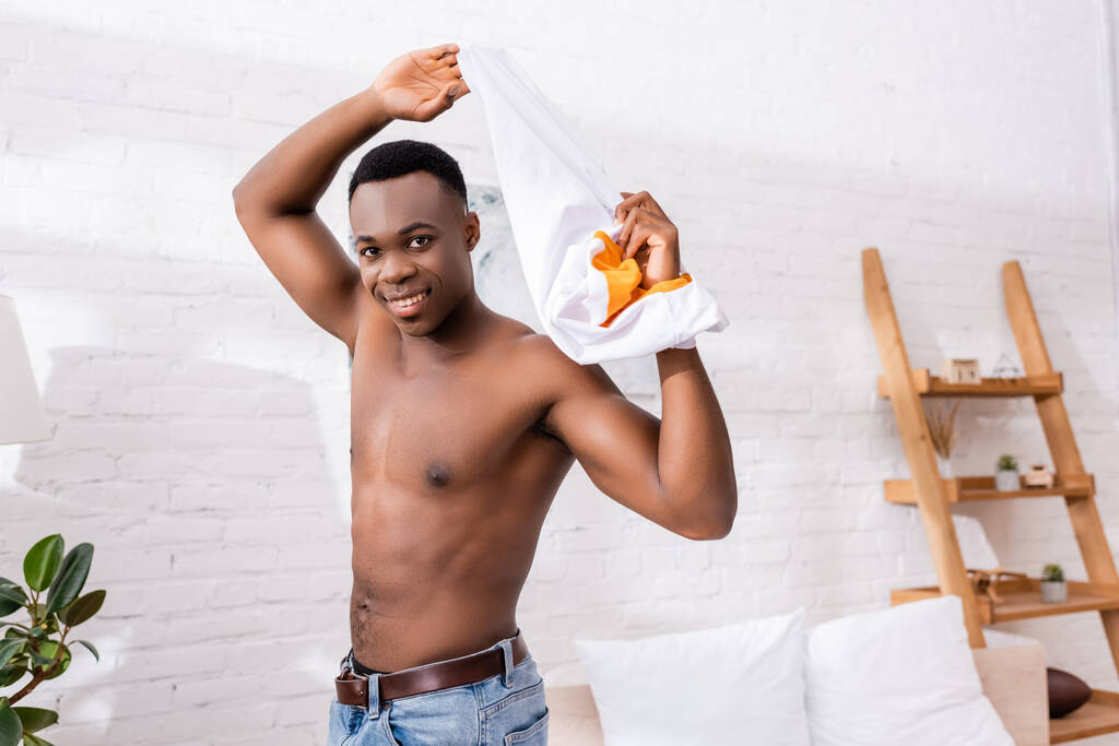 Vrolijke Afro-Amerikaanse man die thuis t-shirt uitdoet  - Foto, afbeelding
