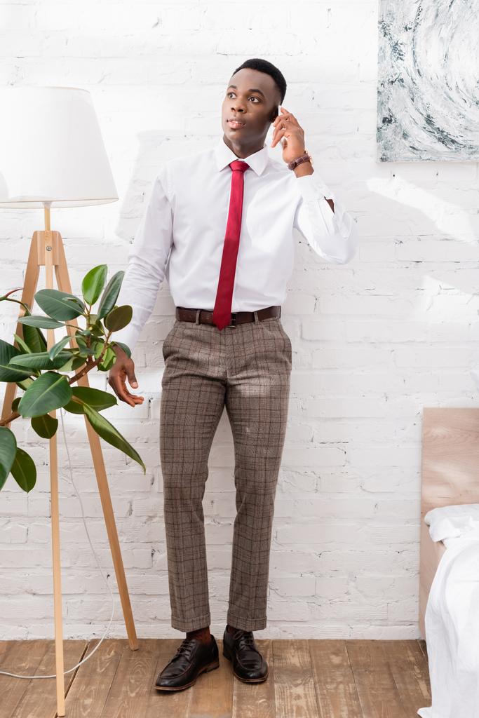 Afrykański amerykański biznesmen rozmawia na smartfonie w pobliżu łóżka i roślin w domu  - Zdjęcie, obraz