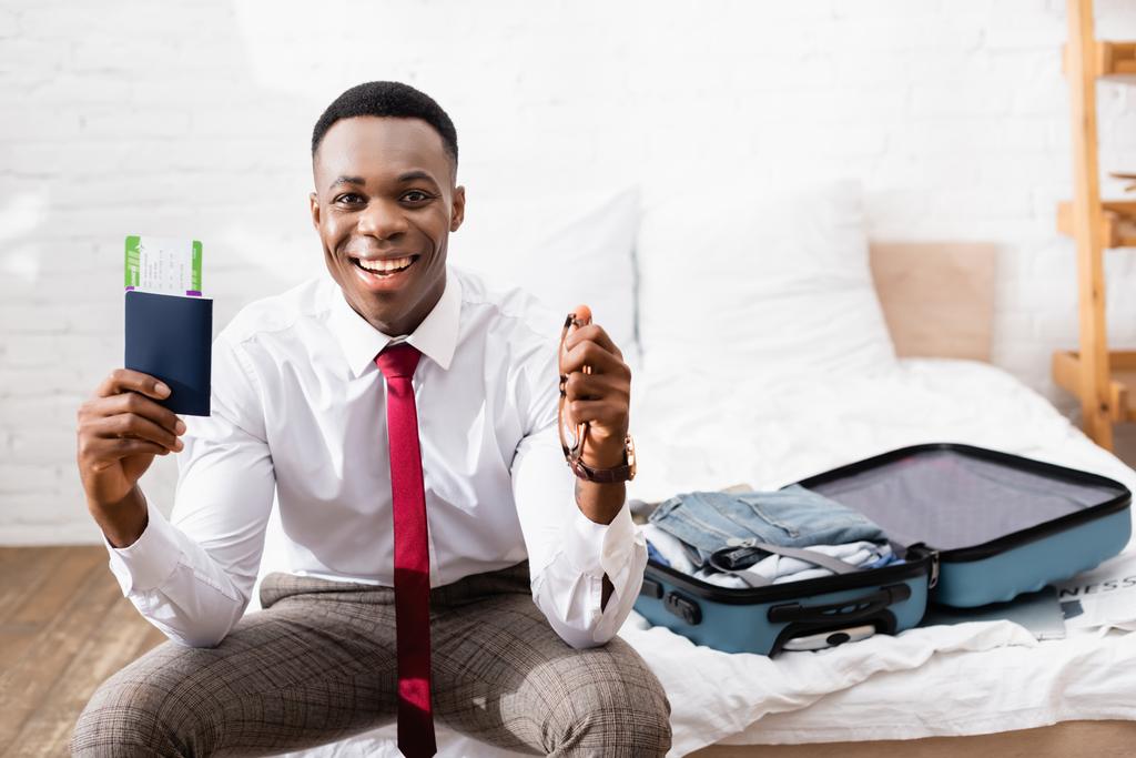 Alegre hombre de negocios afroamericano con pasaporte, billete de avión y anteojos cerca de la maleta sobre fondo borroso en la cama  - Foto, imagen
