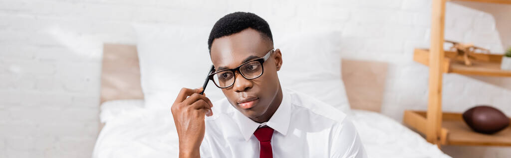 Pensativo hombre de negocios afroamericano en gafas que sostienen la pluma en el dormitorio sobre fondo borroso, pancarta  - Foto, Imagen