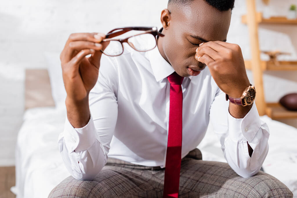 Втомлений афроамериканець, який тримає окуляри на розмитому передньому плані вдома.  - Фото, зображення