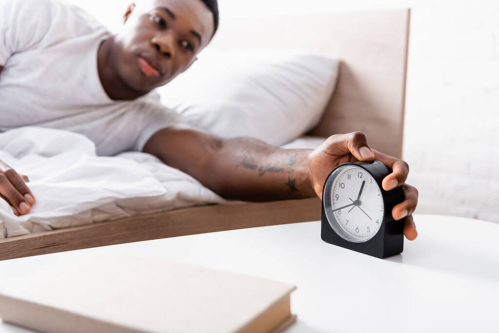 Clock in hand of african american man on bed on blurred background  - Photo, Image