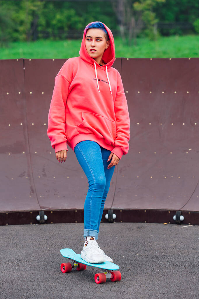 Retrato de uma menina bonita na moda vestida com capuz rosa e jeans de pé ao lado da quadra de skate com seu skate de plástico azul. - Foto, Imagem
