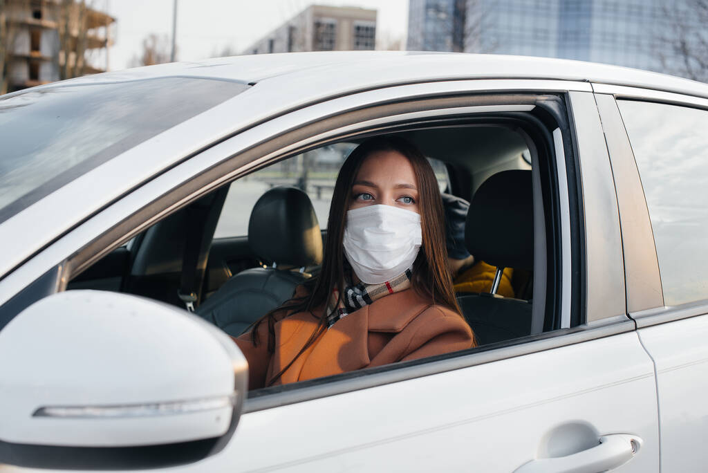 Ein junges Mädchen sitzt während der globalen Pandemie und des Coronavirus hinter dem Steuer des Autos in der Maske. Quarantäne - Foto, Bild