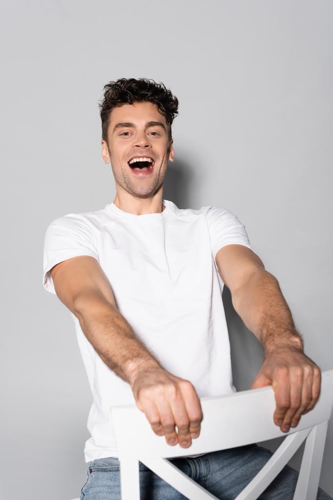 Ευτυχισμένος νεαρός άνδρας με λευκό t-shirt σε καρέκλα απομονωμένη σε γκρι - Φωτογραφία, εικόνα