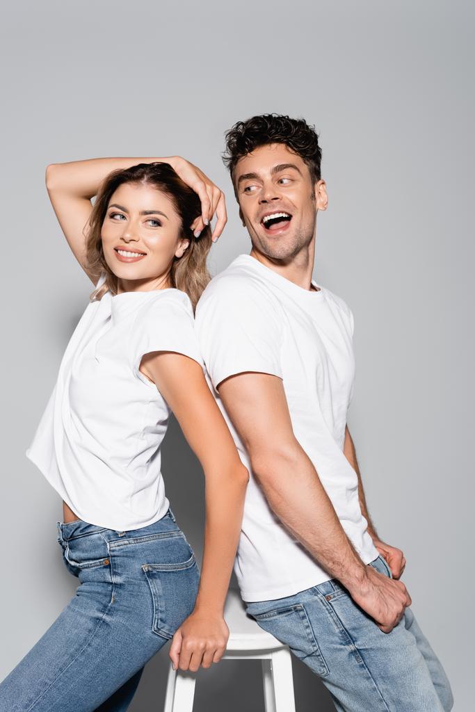 smiling young couple in white t-shirts and jeans posing back to back isolated on grey - Photo, Image