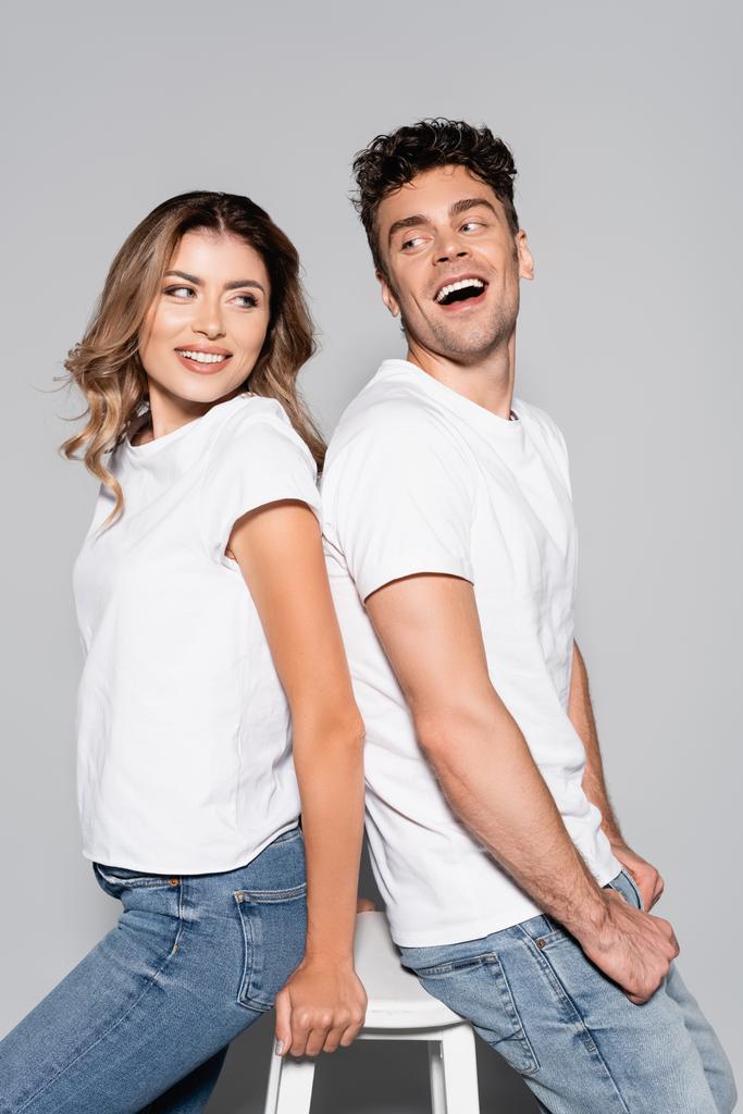 улыбающаяся молодая пара в белых футболках и джинсах, позирующая спина к спине, изолированная на сером - Фото, изображение