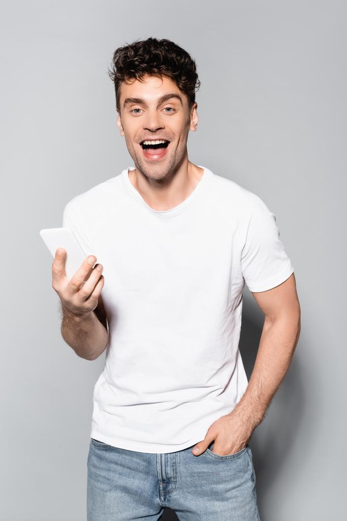onnellinen nuori mies valkoisessa t-paidassa älypuhelimella eristetty harmaalla - Valokuva, kuva