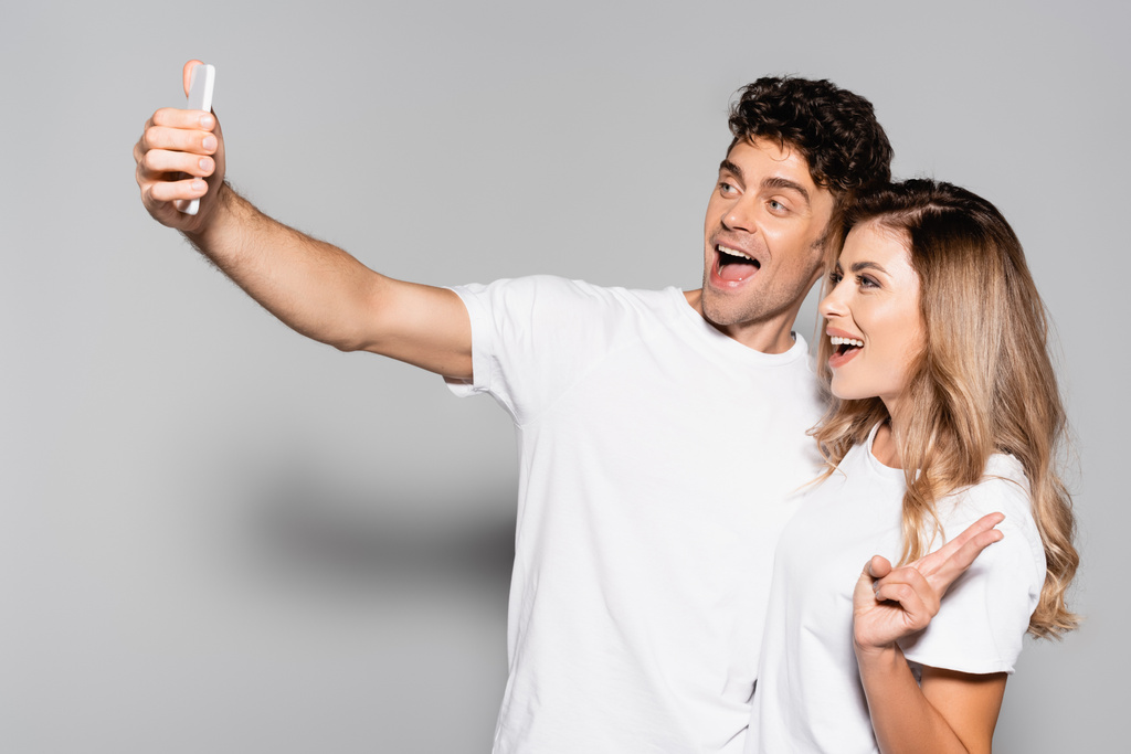 Beyaz tişörtlü mutlu çift, gri renkte izole edilmiş akıllı telefondan selfie çekiyor. - Fotoğraf, Görsel