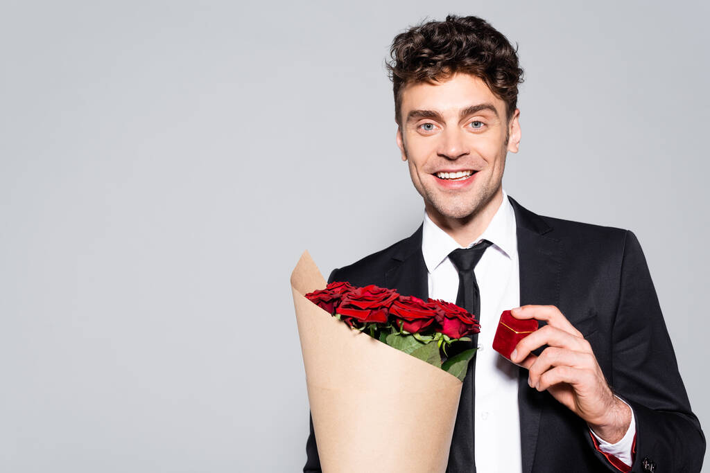 χαμογελαστός άντρας σε κομψό κοστούμι με μπουκέτο κόκκινα τριαντάφυλλα και ring box που απομονώνονται σε γκρι - Φωτογραφία, εικόνα