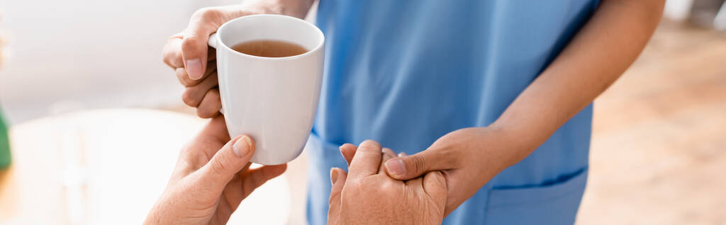 částečný pohled na zdravotní sestru držící se za ruku starší ženy při podávání šálku čaje, rozmazané pozadí, banner - Fotografie, Obrázek