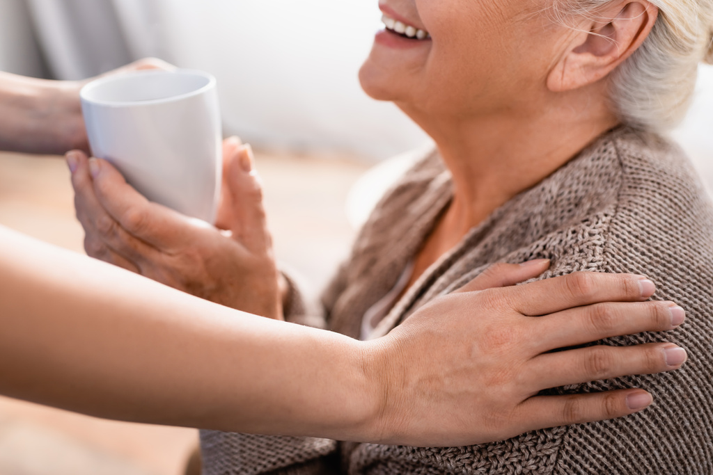 przycięty widok pielęgniarki dotykającej ramienia uśmiechniętej starszej kobiety i dającej filiżankę herbaty, niewyraźne tło - Zdjęcie, obraz
