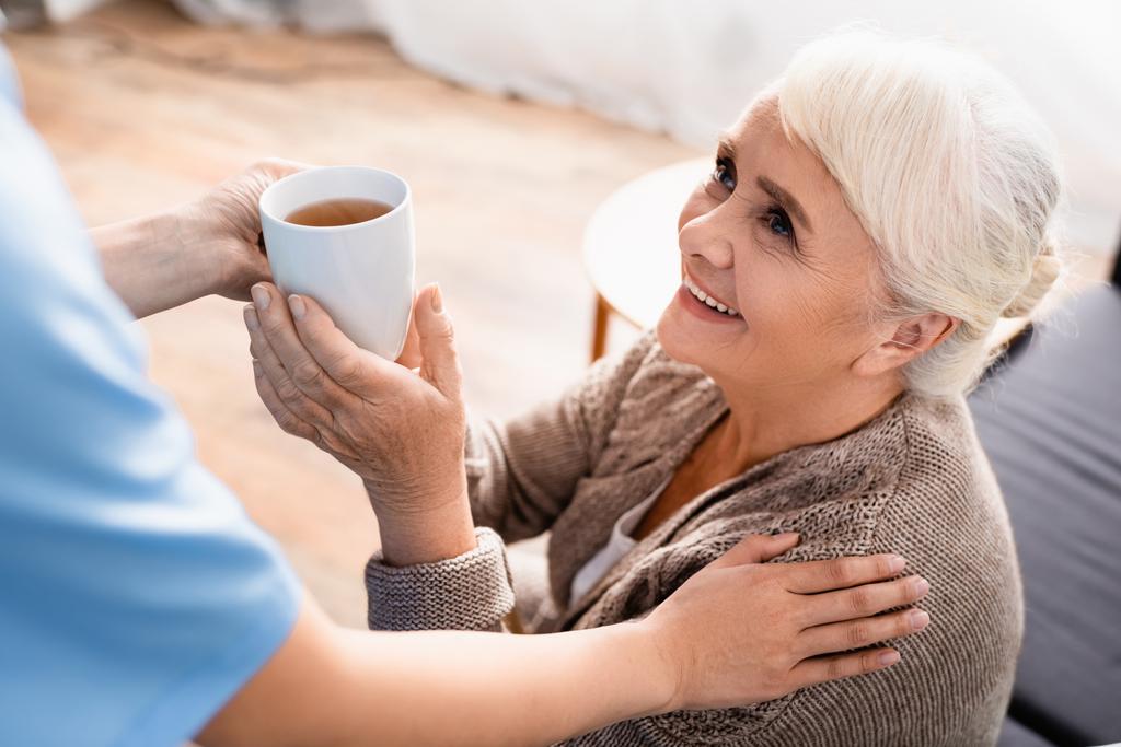 усміхнена старша жінка бере чашку чаю від медсестри на розмитому передньому плані
 - Фото, зображення