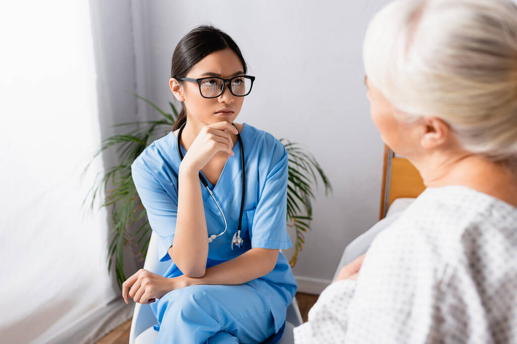 seria asiática enfermera mirando anciana durante conversación en hospital, borrosa primer plano - Foto, imagen