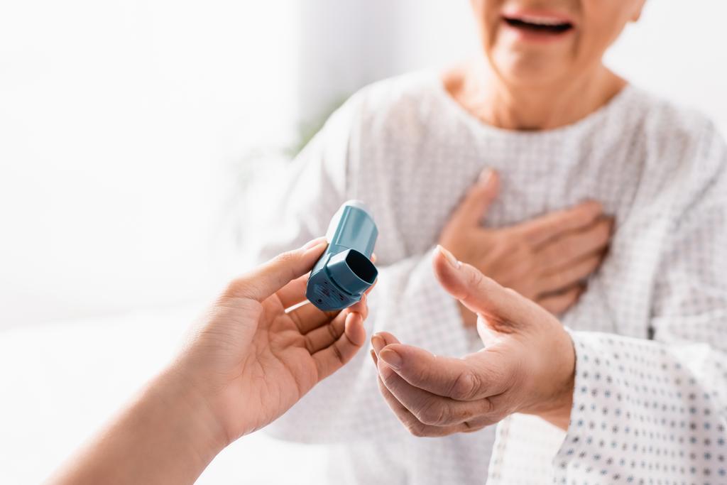 Ausgeschnittene Ansicht einer Krankenschwester, die einer alten Frau, die an Asthmaanfall leidet, vor verschwommenem Hintergrund Inhalator verabreicht - Foto, Bild