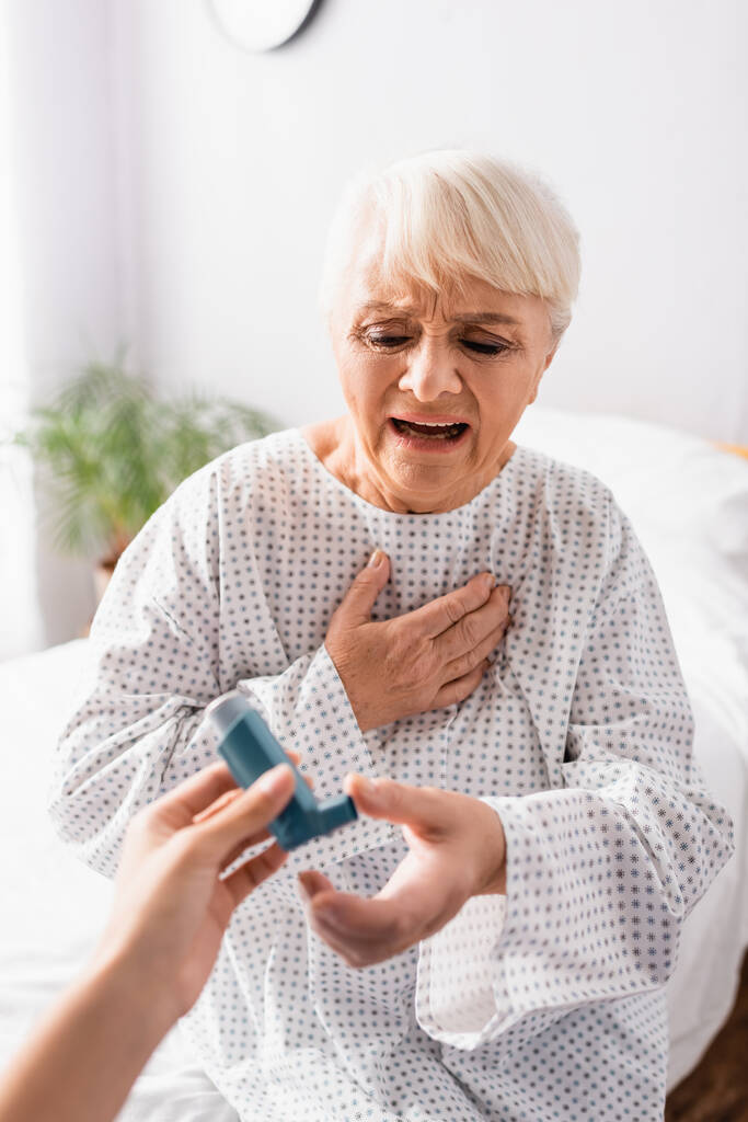 starsza kobieta trzymająca rękę na klatce piersiowej podczas uduszenia i przyjmująca inhalator od pielęgniarki, zamazany pierwszy plan - Zdjęcie, obraz