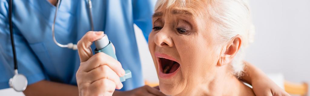 elderly woman with open mouth suffering from asthma attack and using inhaler near nurse on blurred background, banner - Photo, Image